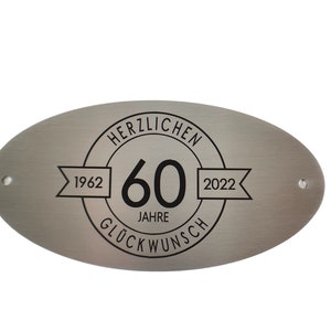 Könnte beinhalten: Ein ovales Schild aus gebürstetem Metall mit einem zentralen kreisförmigen Design, das die Zahl 60, den Text HERZLICHEN GLÜCKWUNSCH sowie die Jahreszahlen 1962 und 2022 auf seitlichen Bannern zeigt. Das Schild hat zwei kleine Befestigungslöcher.