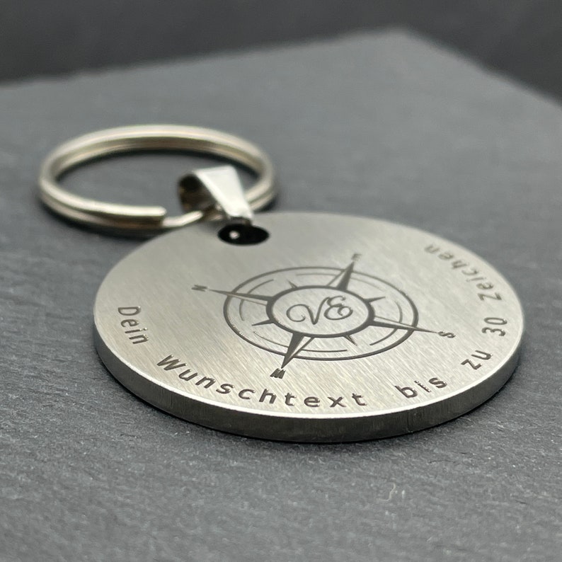 Peut inclure: Un porte-cl&eacute;s en argent avec un motif de boussole et le texte "Dein Wunschtext bis zu 30 Zeichen" grav&eacute; dessus.