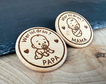 Könnte beinhalten: Zwei Holzmarken mit eingravierten Baby-Designs und dem deutschen Satz "Wer ist dran?" (Wer ist dran?). Eine Marke sagt "PAPA" und die andere sagt "MAMA". Jede Marke hat eine Baby-Illustration und Herz-Akzente.