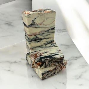 May include: A stack of handmade soap bars with a marbled design. The soap features a creamy white base with swirls of black, brown, and copper tones. The bars are stacked on a white marble surface.