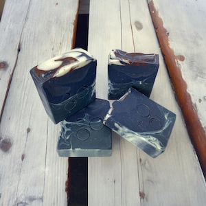 May include: Four bars of handmade soap with a black and white marbled design. The soap is in a rectangular shape and has a white swirl design on top. The soap is sitting on a wooden surface.