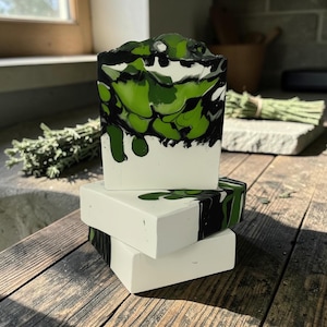 May include: A stack of three rectangular bars of soap. The top bar features a marbled pattern of white, black, and vibrant green colors. The middle bar has a white base with green and black swirls, while the bottom bar is solid white.