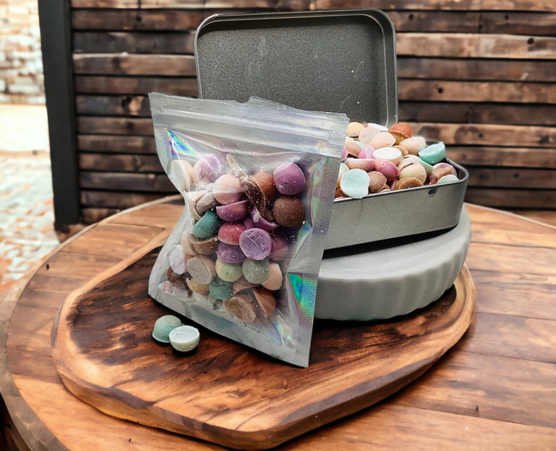 May include: A clear plastic bag filled with colorful round candies, next to a gray metal tin with the same candies inside. The tin is on a wooden surface.