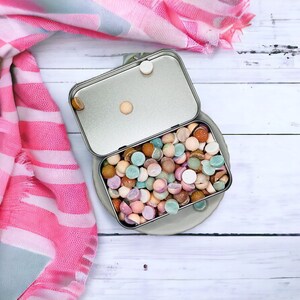 May include: A silver metal tin filled with colorful round candies. The tin is open and the candies are spilling out. The tin is sitting on a white wooden surface.