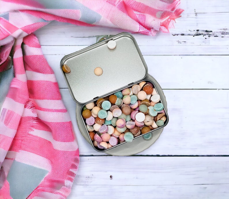 May include: A silver metal tin filled with colorful round candies. The tin is open and the candies are spilling out. The tin is sitting on a white wooden surface.