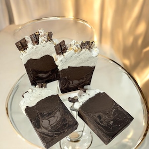 May include: Four bars of handmade soap with a chocolate and marshmallow scent. The soap is swirled with brown and white colors and topped with chocolate pieces and marshmallows.