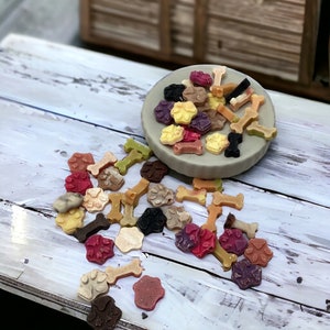 Puede incluir: Una variedad de golosinas para perros de colores en forma de huesos y patas, algunas están en un pequeño tazón blanco, otras están esparcidas sobre una superficie de madera blanca.