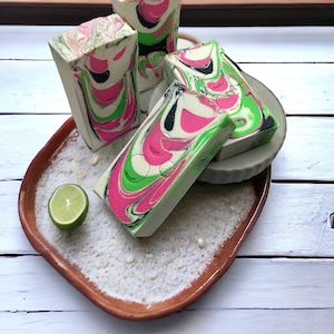May include: Four handmade soap bars with a swirl design in shades of green, pink, and black. The soaps are arranged on a brown ceramic plate with white powder and a lime wedge.