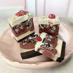 May include: Three handmade soap bars shaped like slices of cake. Each bar has a white layer, a red layer, and a brown layer. The bars are decorated with red and white sprinkles.