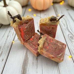 Puede incluir: Cuatro barras de jabón hechas a mano con un diseño en espiral rojo, marrón y beige. La parte superior de cada barra tiene una capa beige texturizada con un pequeño trozo de chocolate negro y una ramita. Los jabones están dispuestos sobre una superficie de madera blanca.