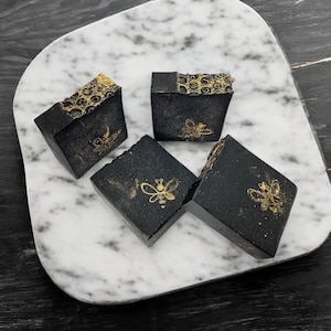 May include: Four black soap bars with gold bee designs on a white marble surface. The soap bars are square shaped and have a honeycomb pattern on the top.