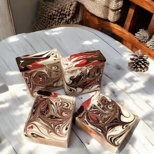 May include: Four handmade soap bars with a swirl pattern in shades of brown, red, and white. The soap bars are arranged on a white wooden surface.