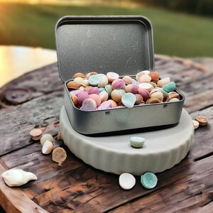 May include: A metal tin filled with colorful round candies, sitting on a wooden surface with a few candies scattered around it.