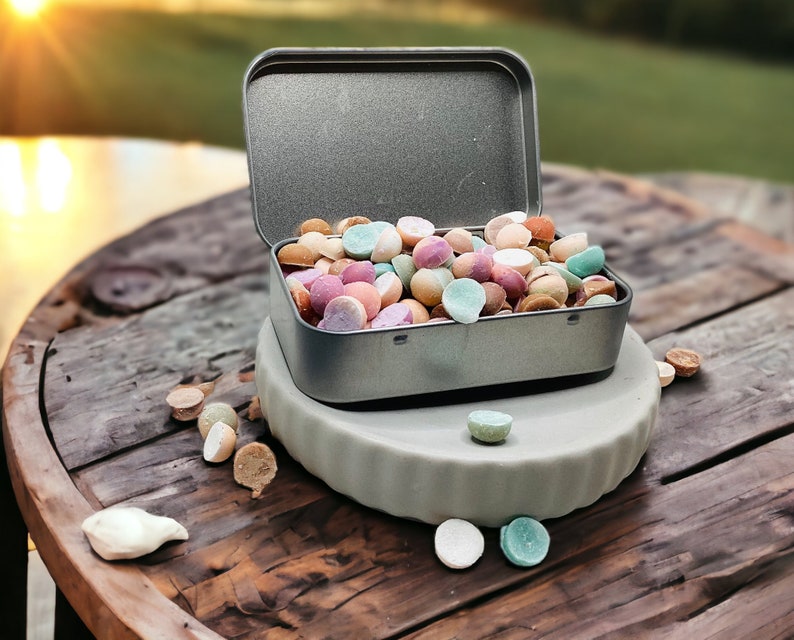 May include: A metal tin filled with colorful round candies, sitting on a wooden surface with a few candies scattered around it.