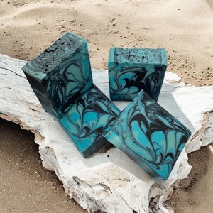 Puede incluir: Cuatro barras cuadradas de jabón artesanal con un diseño de remolino turquesa y negro. El jabón está dispuesto sobre un trozo de madera flotante, con una playa de arena al fondo. El jabón tiene un aspecto veteado.