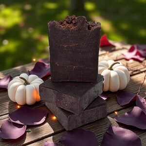 Puede incluir: Jabones rectangulares de color marrón oscuro con una parte superior texturizada, apilados sobre una superficie de madera, con dos pequeñas calabazas blancas y pétalos morados. Los jabones tienen un aspecto natural y artesanal.