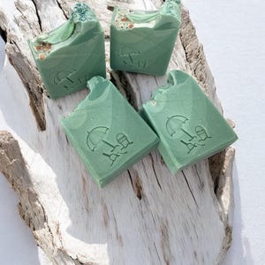 Puede incluir: Cuatro jabones verdes con temática de playa están colocados sobre un trozo de madera flotante. Cada barra tiene un diseño de sombrilla y silla de playa. El jabón tiene una parte superior texturizada con pequeños trozos de color claro.