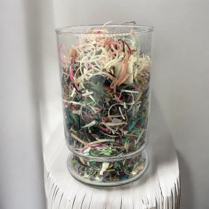 Peut inclure: Un bocal en verre rempli de morceaux de plastique colorés, dont du rose, du vert, du bleu et du blanc. Le bocal est posé sur une surface blanche.