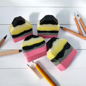 May include: Four handmade soap bars shaped like pencils, with black, yellow, pink, and gray stripes. The soaps are arranged on a white wooden surface with pencils.