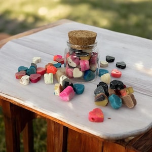May include: A glass jar with a cork lid filled with colorful heart-shaped candies. The jar is sitting on a white wooden table with more candies scattered around it.