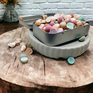 May include: A metal tin filled with colorful candy, sitting on a wooden surface. The candy is in the shape of small, round balls and is a variety of colors, including pink, green, yellow, and orange.