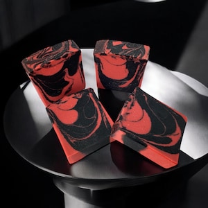 May include: Four bars of handmade soap with a red and black marbled design. The soap is displayed on a black surface.