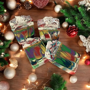 May include: Four colorful, handmade soap bars with a marbled design, featuring shades of red, green, purple, and yellow. The soaps have a textured, white top and are arranged on a wooden surface with Christmas ornaments and greenery.