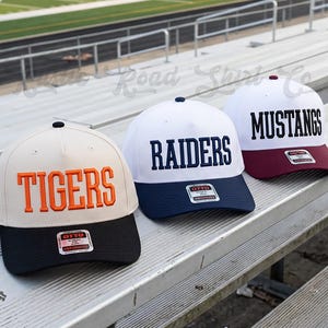 May include: Three baseball caps with structured crowns and flat brims. The first is cream with orange TIGERS text and a black brim. The second is white with navy RAIDERS text and a navy brim. The third is white with black MUSTANGS text and a maroon brim.