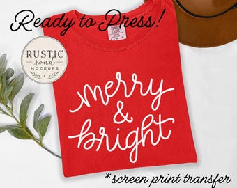 May include: A red t-shirt featuring the white cursive text merry & bright centered on the front. The shirt has a Comfort Colors brand label at the neckline. The text Ready to Press! and screen print transfer are visible in the image.