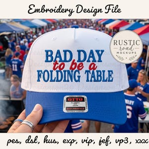May include: A white and blue baseball cap featuring the embroidered text "BAD DAY to be a FOLDING TABLE" in blue and red. The cap includes a sticker on the brim with the text "OTTO CLASSIC" and "MID PROFILE" and is presented as an embroidery design file.