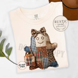 May include: A cream-coloured t-shirt featuring a graphic of a patchwork ghost figure with a leopard print bow, carrying a small bag with a pumpkin. The shirt has a Comfort Colors label at the neck and a circular logo reading Rustic Road Mockups.