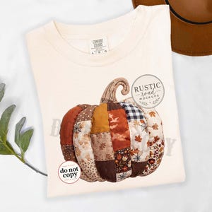 May include: A cream-colored t-shirt featuring a large graphic of a patchwork pumpkin. The pumpkin is composed of various fabric patterns in shades of orange, brown, and black, including leaves, plaid, and spiderwebs. Text reads Rustic Road Mockups.