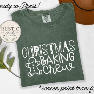 Puede incluir: Una camiseta de color verde oliva oscuro con el texto en blanco "CHRISTMAS BAKING CREW" y un gráfico de un hombre de jengibre. La prenda tiene un cuello redondo y una etiqueta de la marca Comfort Colors visible en el interior.