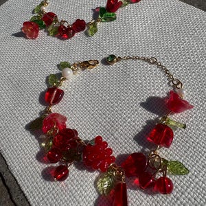 May include: Two gold-tone chain bracelets featuring translucent red glass charms shaped like flowers, berries, and cherries, accented with small green glass leaves and white spherical beads. Each bracelet includes a lobster claw clasp closure.