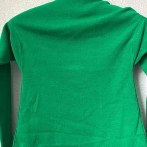 May include: A vibrant green turtleneck jumper with long sleeves. The jumper is made of a ribbed knit fabric and is hanging on a wooden hanger. The jumper's bright colour and classic design make it a versatile wardrobe staple.