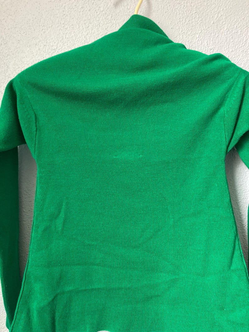 May include: A vibrant green turtleneck jumper with long sleeves. The jumper is made of a ribbed knit fabric and is hanging on a wooden hanger. The jumper's bright colour and classic design make it a versatile wardrobe staple.