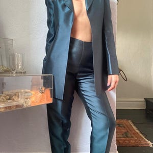 May include: Teal-colored satin blazer and matching pants. The blazer has a classic lapel and a tailored fit. The pants are high-waisted with a straight leg. The outfit is made of a shiny, luxurious fabric.