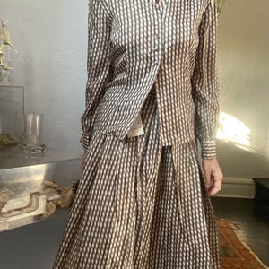 May include: A two-piece vintage outfit featuring a button-down shirt and a pleated skirt. The set is patterned with vertical stripes in shades of brown and cream. The shirt has long sleeves and a collared neckline. The skirt falls to mid-calf length.