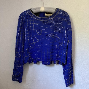 May include: A royal blue long-sleeve top with silver embellishments. The top features a grid pattern with heart and rectangular designs. The neckline and cuffs are trimmed with silver. The garment is hanging on a hanger.