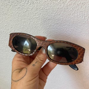 May include: Oval sunglasses featuring a brown and black snakeskin-patterned frame with dark tinted lenses. The frame has a textured, scale-like appearance, and the temples are black. These eyewear pieces provide a distinct look with their patterned finish.