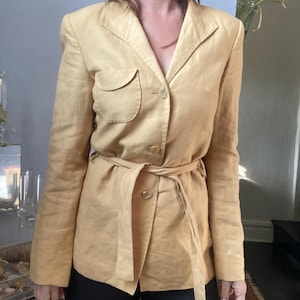 May include: A tan blazer with a belted waist and button closure. The blazer features a notched collar, a breast pocket, and long sleeves. The fabric appears to be a lightweight material, suitable for warmer weather. The blazer is a neutral color.
