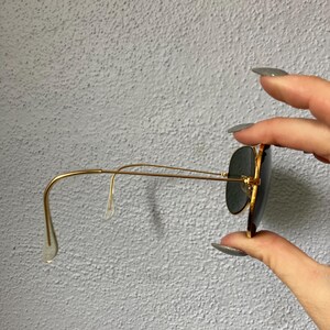 Op de afbeelding: Goudkleurige zonnebril met donkere glazen. De zonnebril heeft een klassieke pilotenstijl met een dubbele brug en een dun goudkleurig montuur. De pootjes buigen naar de oren toe. De glazen zijn donker van kleur.