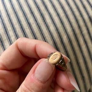 May include: A gold-toned metal ring featuring an oval-shaped face with an engraved eye design. The band of the ring displays a decorative, open-work grid pattern on the sides, extending from the central oval setting to the rest of the band.