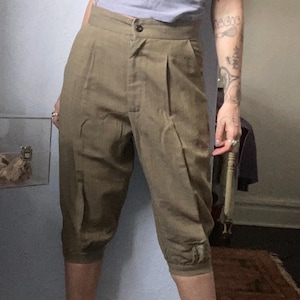 May include: Khaki-colored, knee-length pants with a button closure and pleated front. The pants have a tailored fit and are made of a woven fabric. The cuffs are gathered at the bottom. The pants are worn by a person.