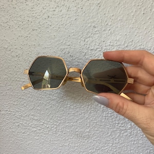 May include: Gold-framed sunglasses with a unique hexagonal shape. The lenses are a dark gray color, and the frames have a textured design on the temples. The sunglasses are held against a light gray wall.