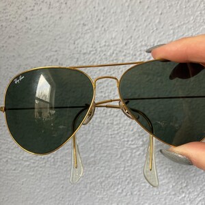 Op de afbeelding: Goudkleurige piloten zonnebril met donkergroene glazen. Het Ray-Ban logo is zichtbaar op het linkerbovenglas. De zonnebril heeft transparante neuspads.
