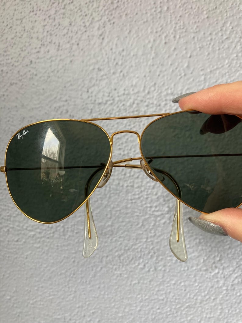 Op de afbeelding: Goudkleurige piloten zonnebril met donkergroene glazen. Het Ray-Ban logo is zichtbaar op het linkerbovenglas. De zonnebril heeft transparante neuspads.