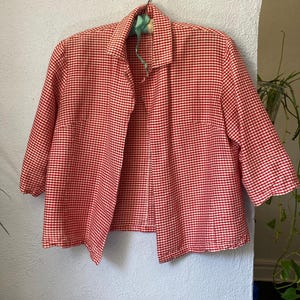 May include: A red and white gingham patterned jacket with a collared neckline and three-quarter length sleeves. The garment features a buttonless open front design and a straight hemline, showcasing a classic checkered textile pattern throughout.