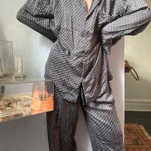 May include: A two-piece pajama set consisting of a long-sleeved button-down shirt and matching loose-fitting trousers. The set is dark gray with a repeating white geometric pattern of small squares and dots throughout the fabric.