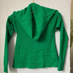 May include: A vibrant green sweater with a cowl neck, hanging on a wooden hanger against a white wall. The long-sleeved sweater features a ribbed texture and a folded cowl neckline. The garment is made of a soft, knitted material.